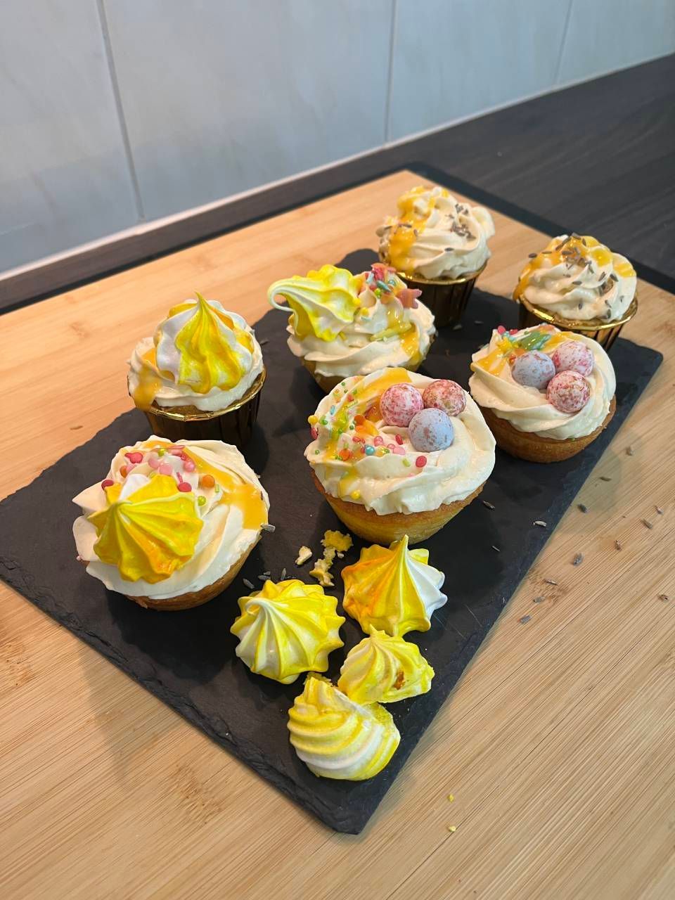 Verschiedene glasierte Cupcakes auf einem schwarzen Tablett, mit gelbem, rosa und weissem Zuckerguss auf einem Holztisch.