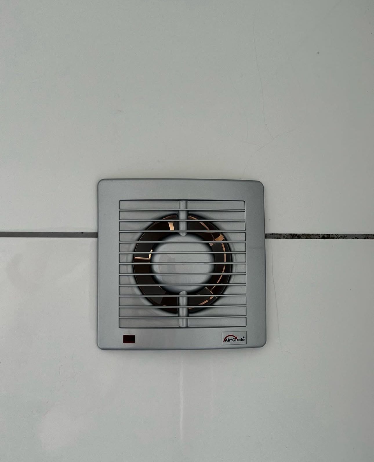An einer weiß gefliesten Wand hängt ein Ventilator.