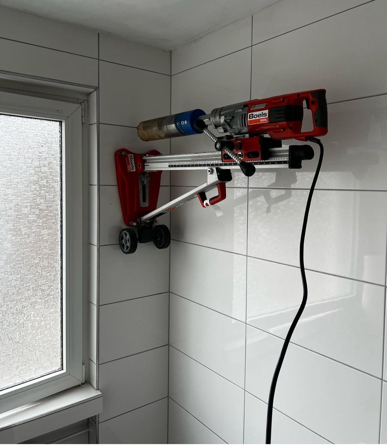An einer weiß gefliesten Wand neben einem Fenster hängt eine rote Bohrmaschine.