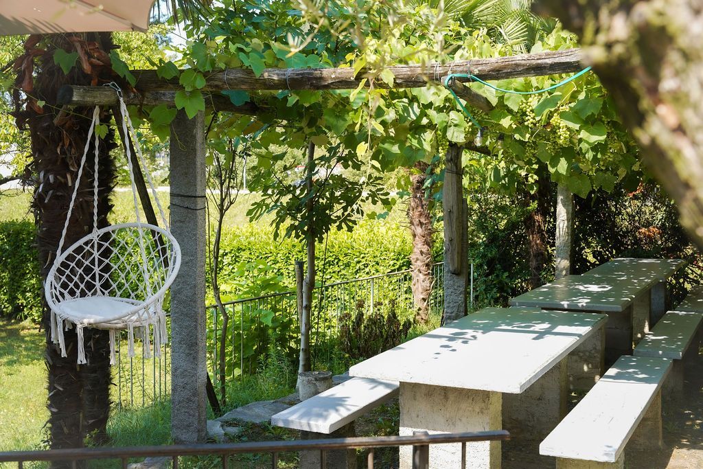 Patio in giardino con lungo tavolo bianco e panchine sotto un pergolato ricoperto di piante rampicanti, con una sedia sospesa bianca.