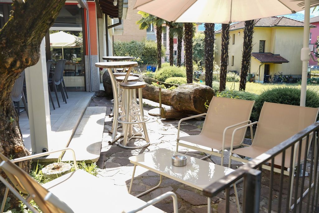 Patio esterno con tavoli, sedie, ombrelloni e piante in vaso accanto a un edificio