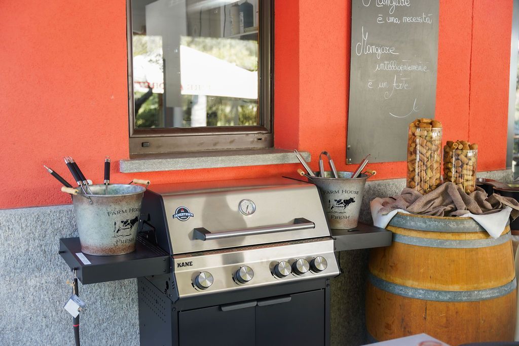 Area barbecue esterna con barbecue argentato, piante in vaso e una botte di legno contro un muro rosso.