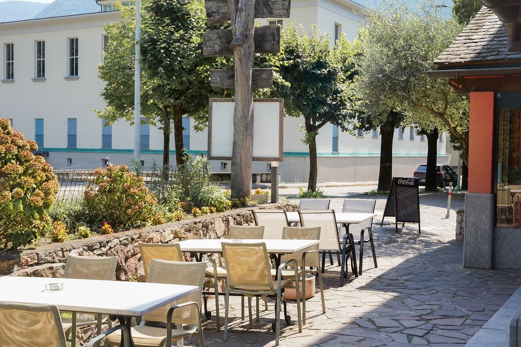 Tavolini all'aperto di un caffè su una soleggiata strada acciottolata, accanto a un edificio bianco e agli alberi.