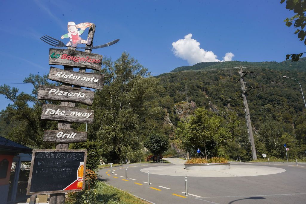 Cartello stradale a lato di una strada di montagna sotto un cielo azzurro con alberi e colline