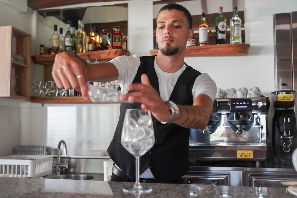Un barista versa un drink in un bicchiere in cucina, con uno shaker e delle bottiglie sul bancone.