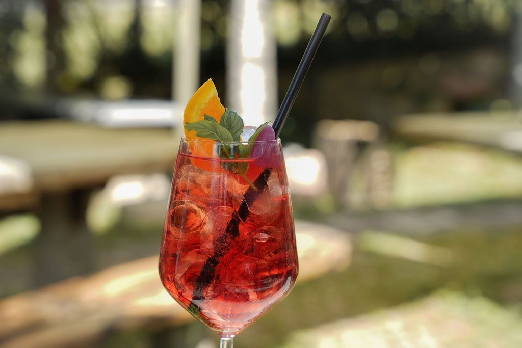 Cocktail rosso in un bicchiere con stelo, guarnito con arancia e cannuccia nera, servito all'aperto.