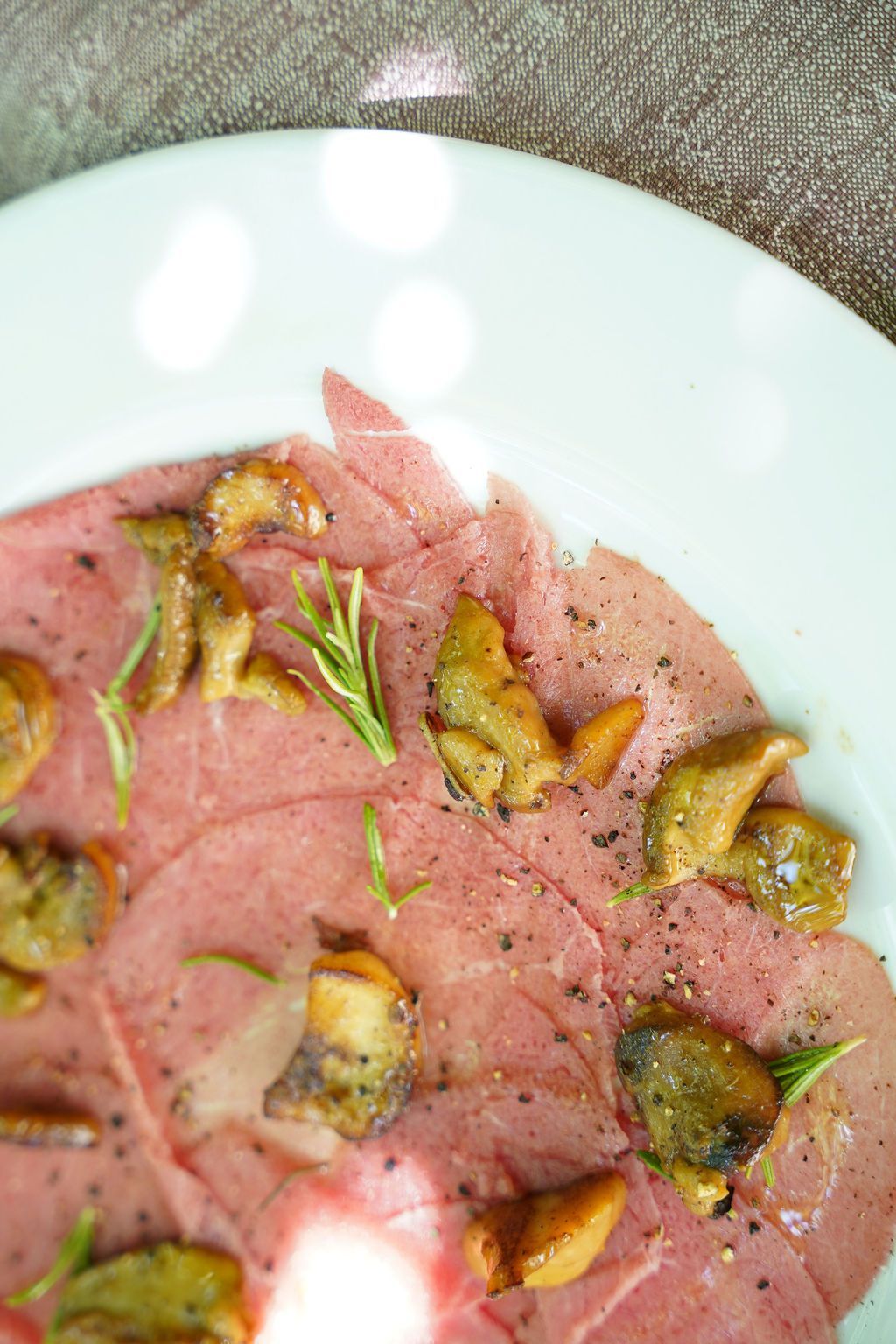 Antipasto di salumi rosati con funghi rosolati e rosmarino, servito su un piatto bianco.