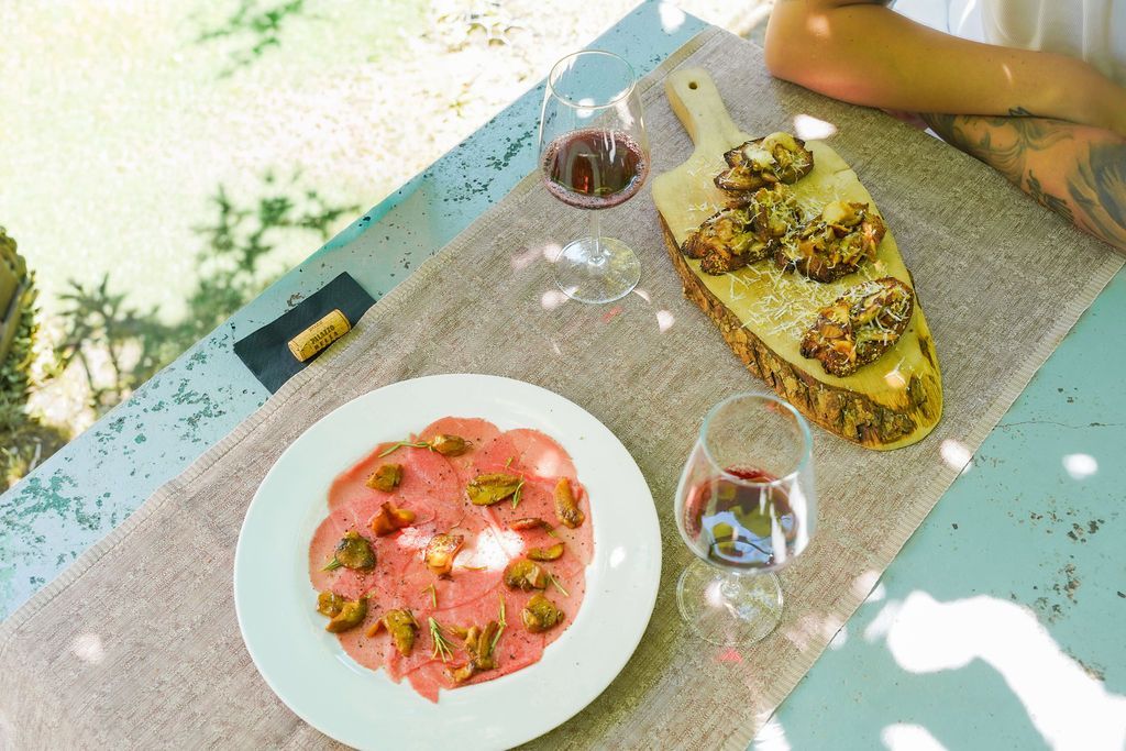 Un tavolo con pasta rosa, una focaccia condita e due bicchieri su un patio soleggiato.