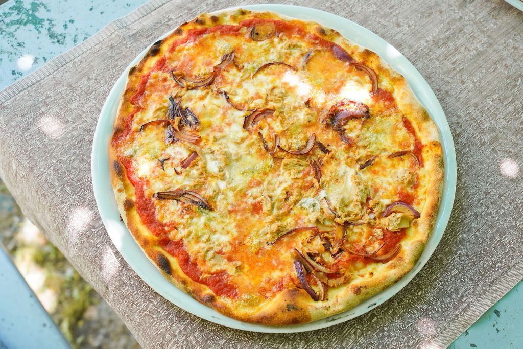 Una pizza rotonda al formaggio su un piatto azzurro chiaro all'aperto, su una tovaglia dalla trama particolare.