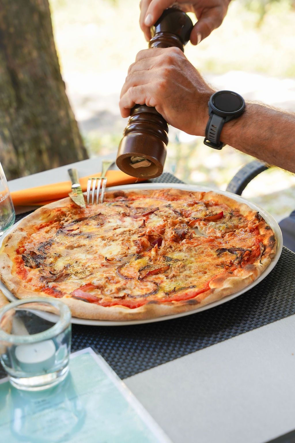 Condire a mano una pizza al formaggio a un tavolo all'aperto con bevande e uno smartwatch