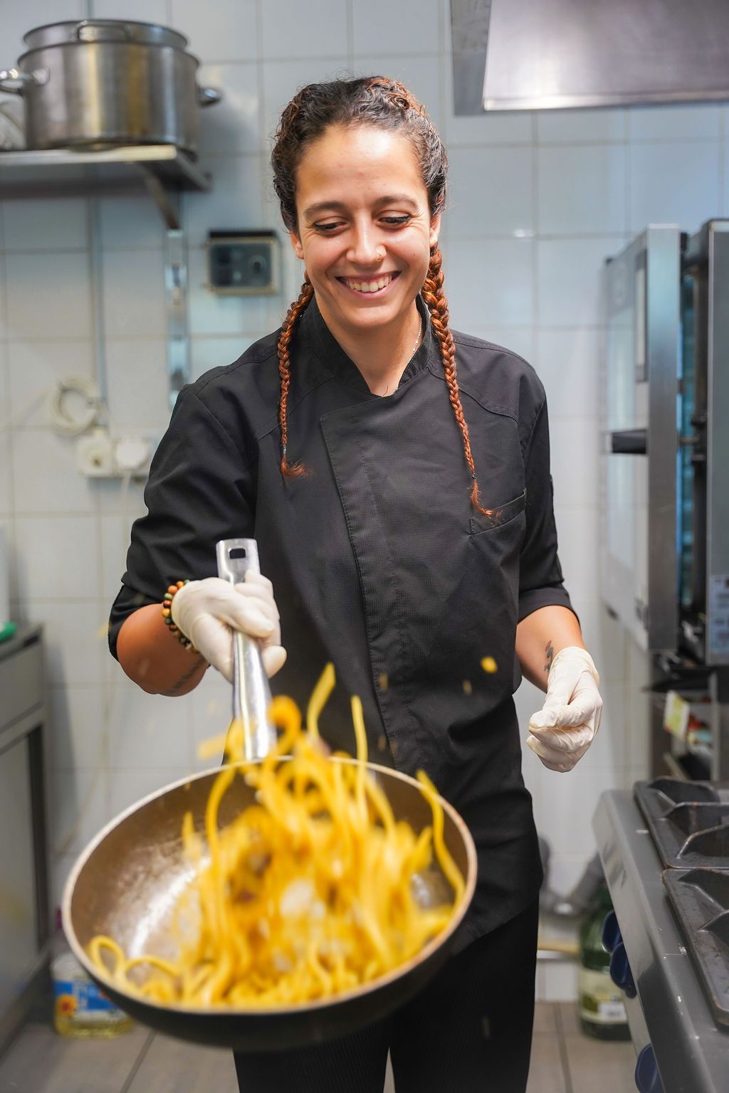 Uno chef sorridente, in uniforme nera, salta la pasta in padella in una cucina professionale.