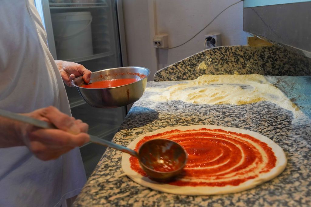 In cucina, una persona spalma la salsa rossa sull'impasto della pizza con un mestolo.