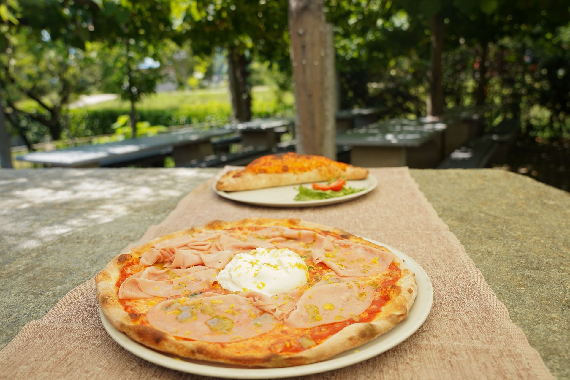 Piatto di pizza con prosciutto crudo su un letto di verdure con pomodorini, servito all'aperto su un tavolo di pietra.