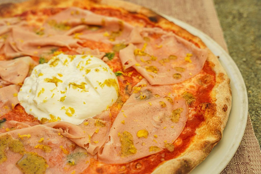 Pizza condita con fette di prosciutto, un uovo in camicia e origano su un tavolo di legno.