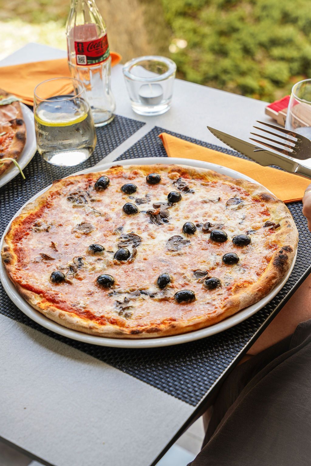 Pizza su un tavolo all'aperto con bevande, tovaglioli e posate in un ambiente informale.