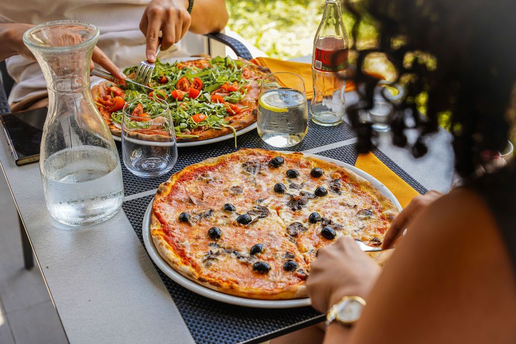 Tavolo all'aperto con pizze, insalata, bevande e persone che si servono da sole.