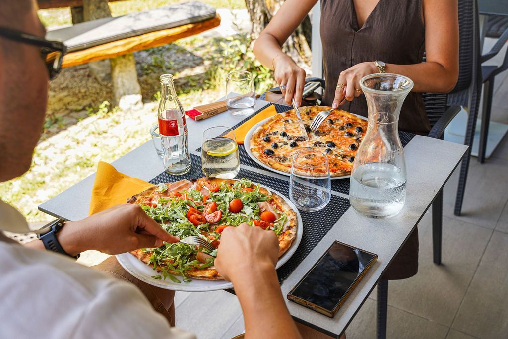 Due persone che condividono una pizza a un tavolino di un bar, con una brocca di vetro e uno smartphone nelle vicinanze.