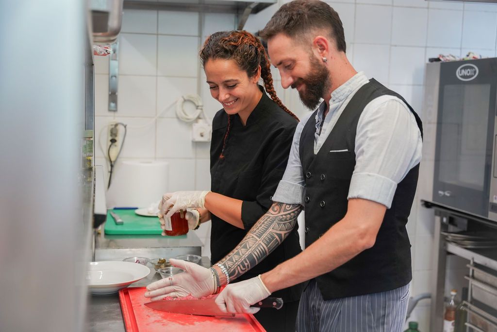 Due persone, con indosso grembiuli e guanti, preparano del cibo su un bancone della cucina, con un'espressione sorridente.