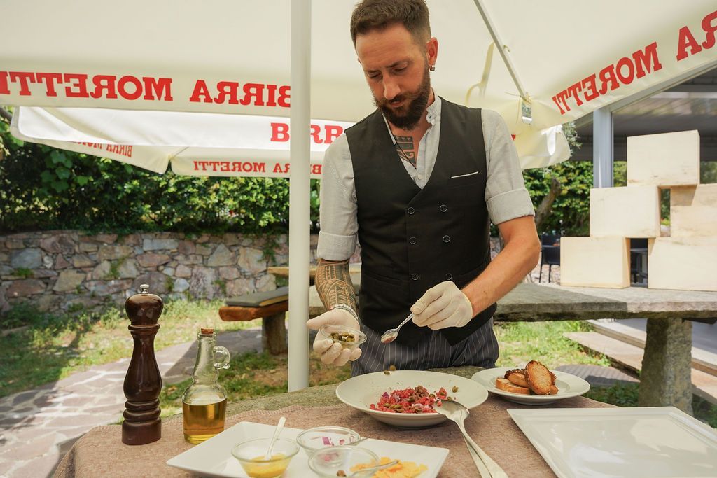 Lo chef impiatta le pietanze su un tavolo all'aperto, con salse e vino, in un ambiente da giardino.