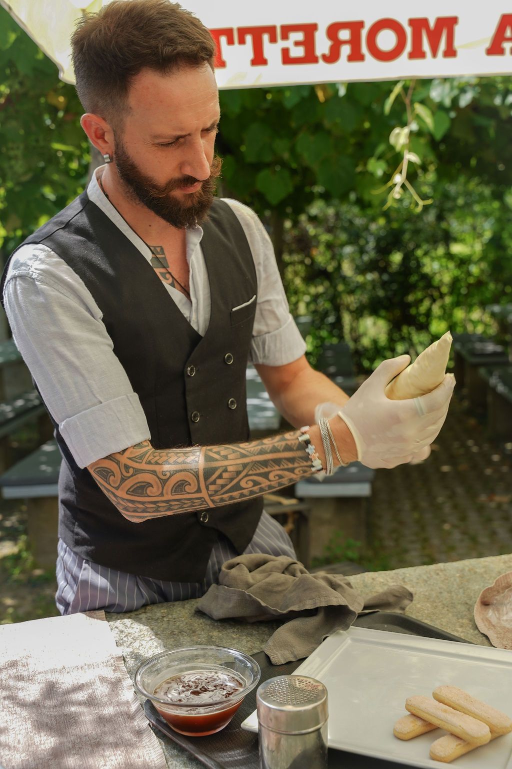 Un uomo con gilet e guanti prepara del cibo presso una griglia all'aperto, con salsa e pane nelle vicinanze.