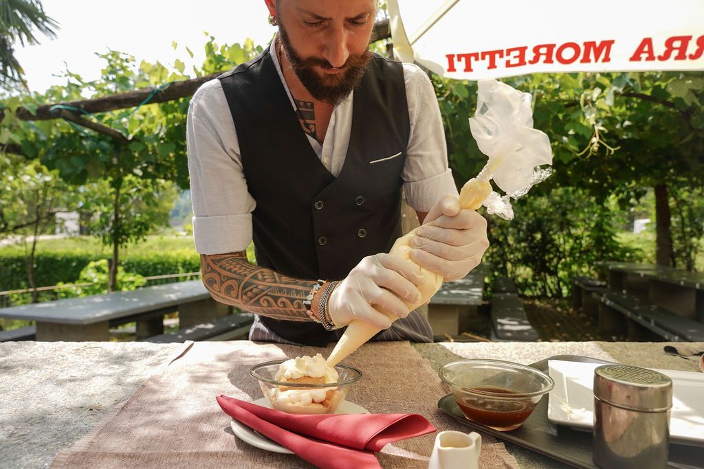 Lo chef decora un dessert con panna bianca all'aperto, seduto a un tavolo con ciotole e utensili.