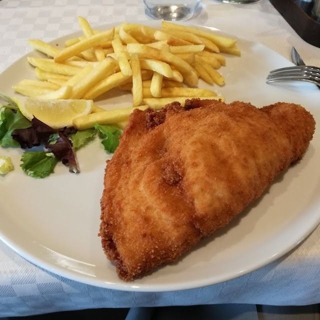 Piatto con cotoletta fritta impanata, patatine fritte e una piccola insalata verde su una tovaglia bianca.