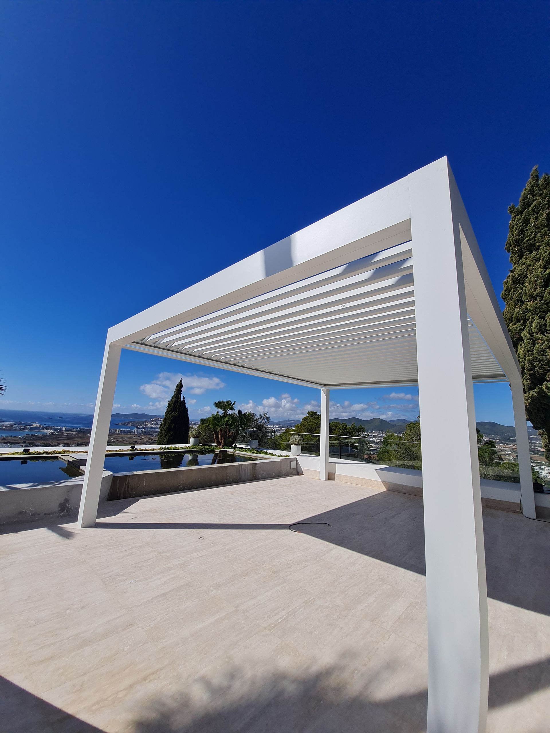 Pérgola blanca en una terraza soleada con sombras rayadas bajo un cielo azul claro.