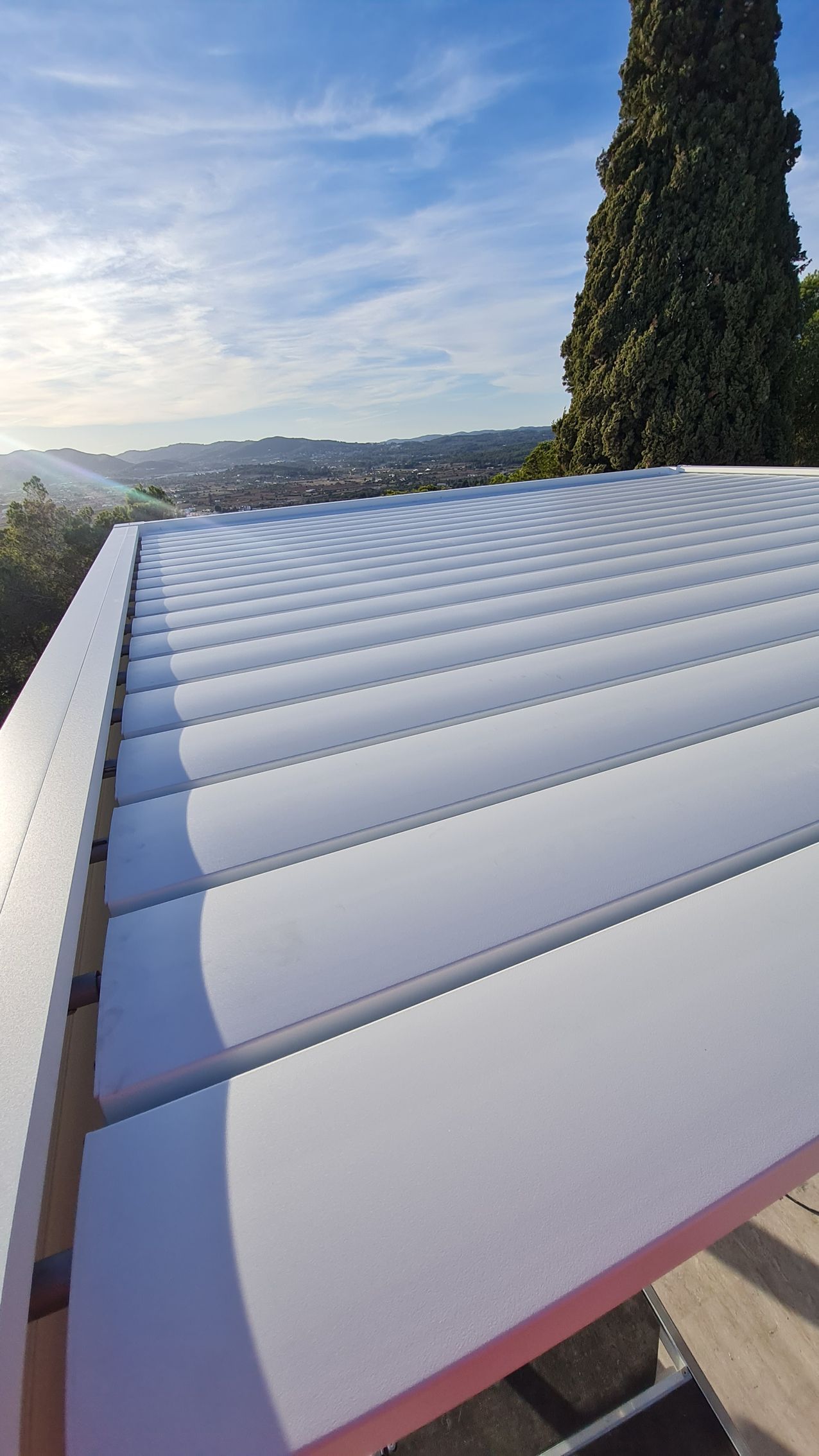 Pérgola con techo de listones blancos, cielo azul y colinas lejanas más allá de una terraza en la ladera.