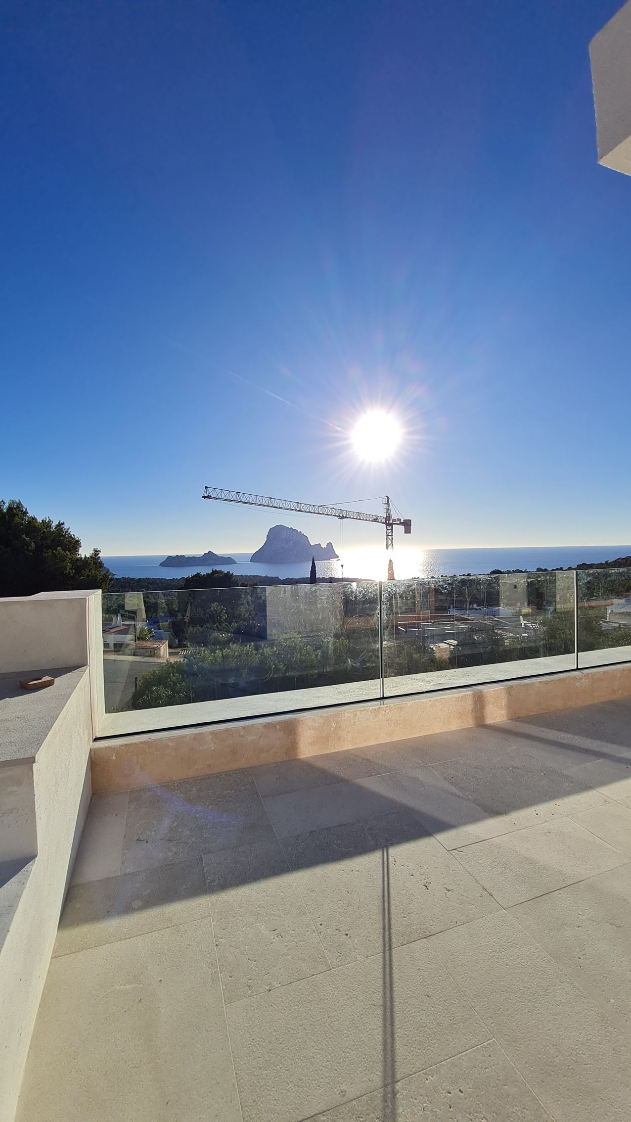 Terraza soleada en la azotea con barandilla de cristal, cielo azul y vistas lejanas al mar y a los edificios al atardecer.