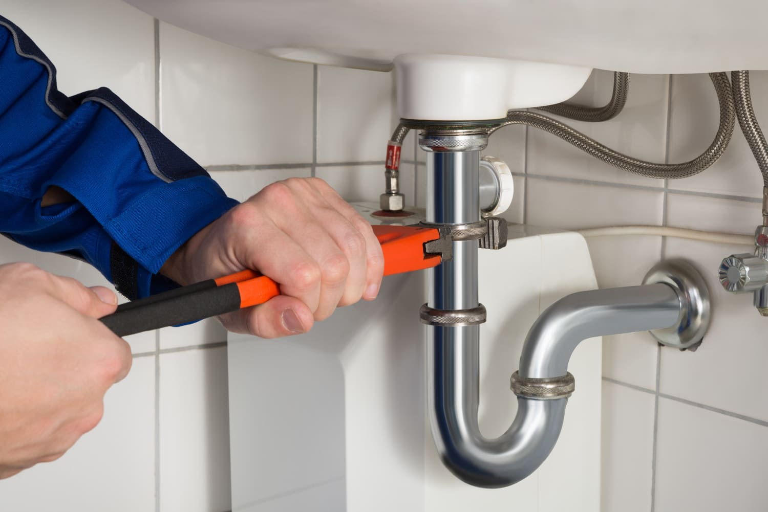 Un fontanero con uniforme azul utiliza una llave inglesa para apretar una tubería debajo de un lavabo blanco en un baño.