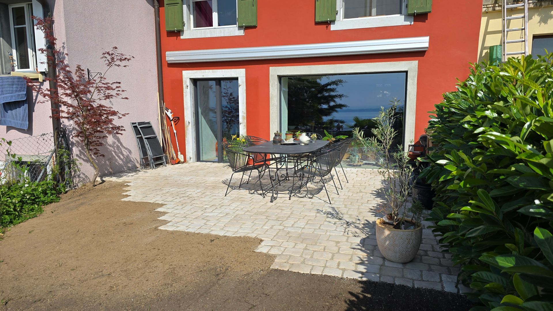 Terrasse d'une maison rouge avec table et chaises d'extérieur, allée de gravier et arbustes de jardin verts.