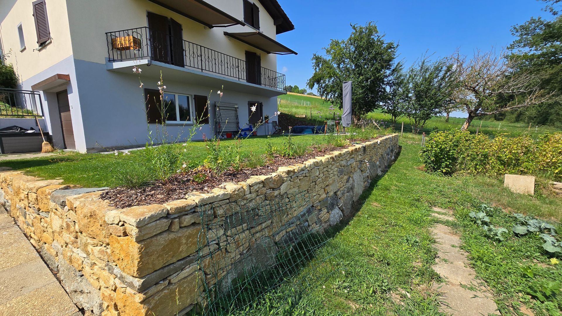 Maison avec balcon jouxtant un mur de soutènement en pierre et une cour gazonnée sous un ciel d'un bleu éclatant