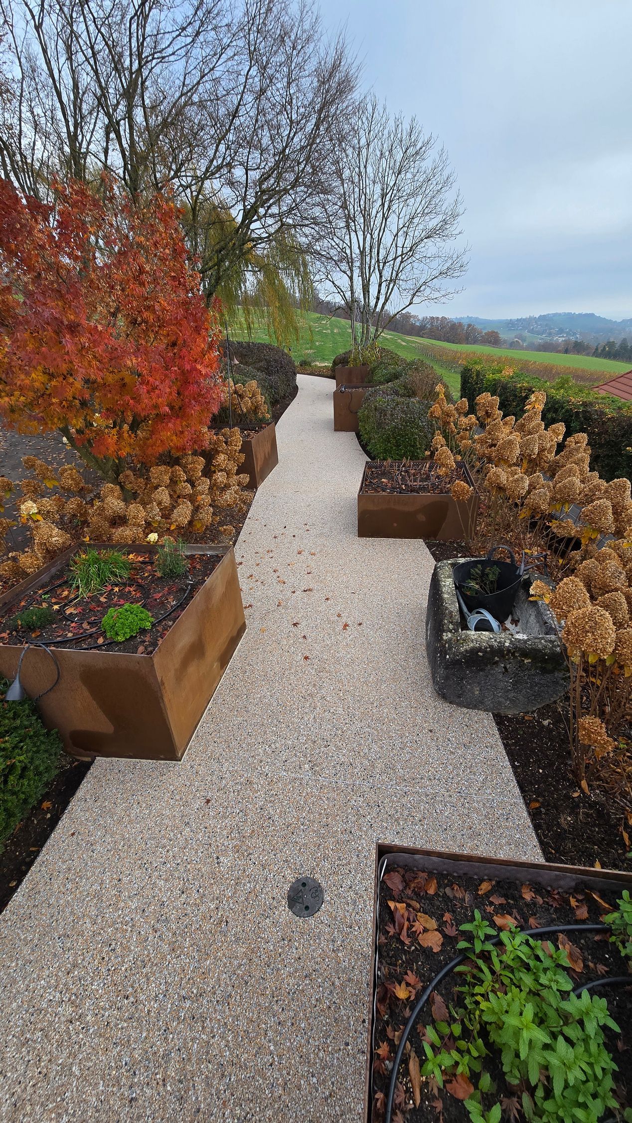 Chemin de gravier traversant des parterres de jardin colorés, avec des feuillages d'automne et des collines au loin.