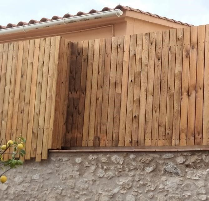 Una valla de madera rodea un muro de piedra frente a una casa.