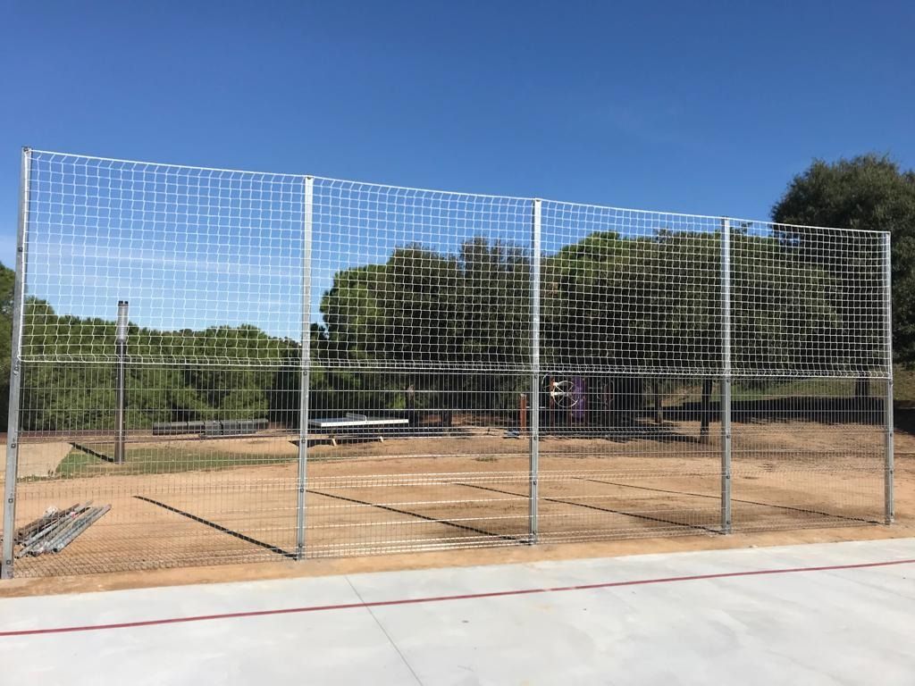 Una valla metálica rodea un campo de béisbol con árboles al fondo.