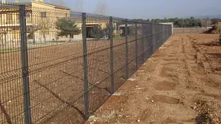 Una valla rodea un campo de tierra con una casa al fondo.