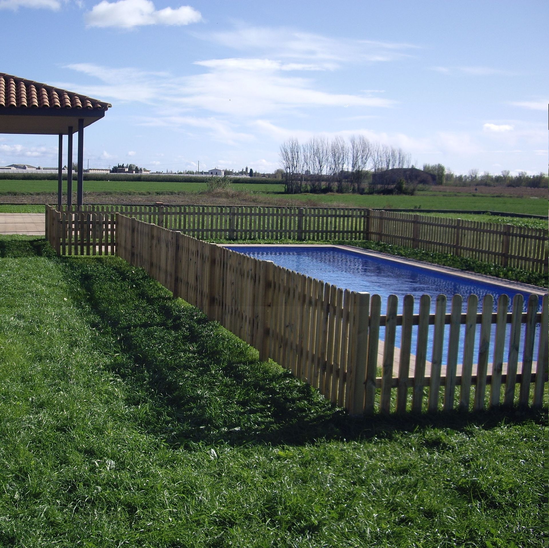 Una valla de madera rodea una piscina con un cenador al fondo.