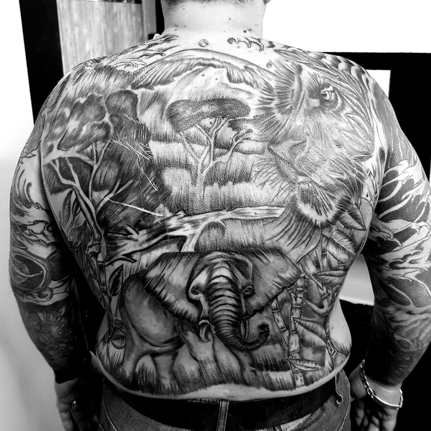 Foto en blanco y negro de la espalda de un hombre cubierta de tatuajes detallados, entre ellos elefantes y un tigre.