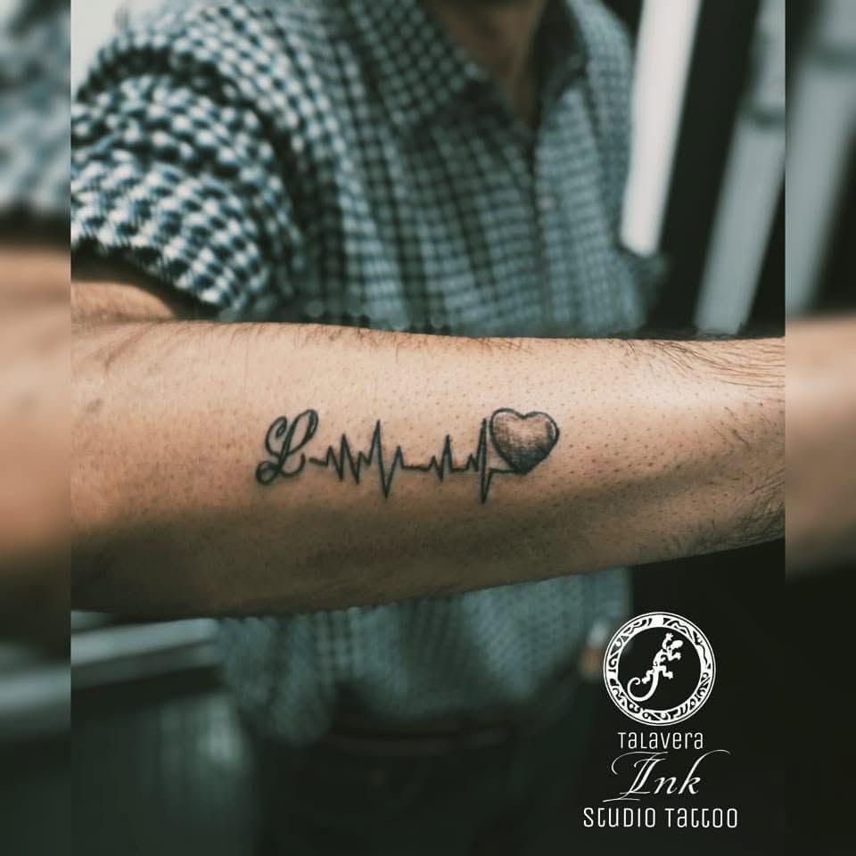 Antebrazo con tatuaje de un corazón y un latido, camisa de cuadros desenfocada al fondo