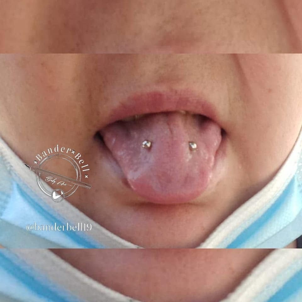 Primer plano de una boca abierta con la lengua extendida y dos pequeños piercings metálicos en la lengua.