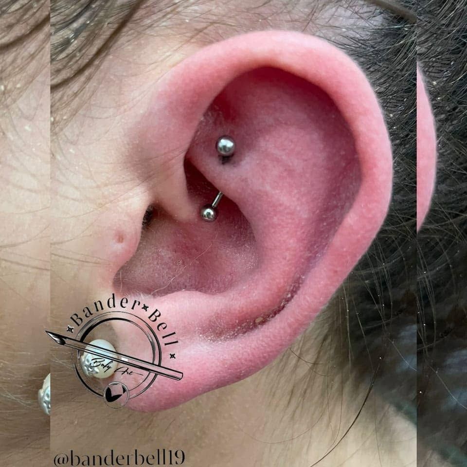 Primer plano de una oreja perforada con una barra curva plateada y un pendiente de botón, junto a una pegatina de un pendiente de dibujos animados.