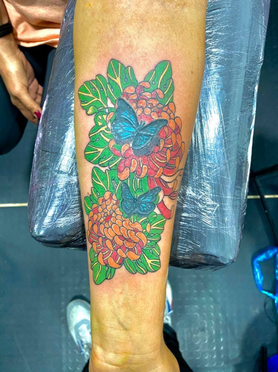 Tatuaje colorido de una mariposa entre flores en el antebrazo de una persona, con hojas verdes y flores naranjas