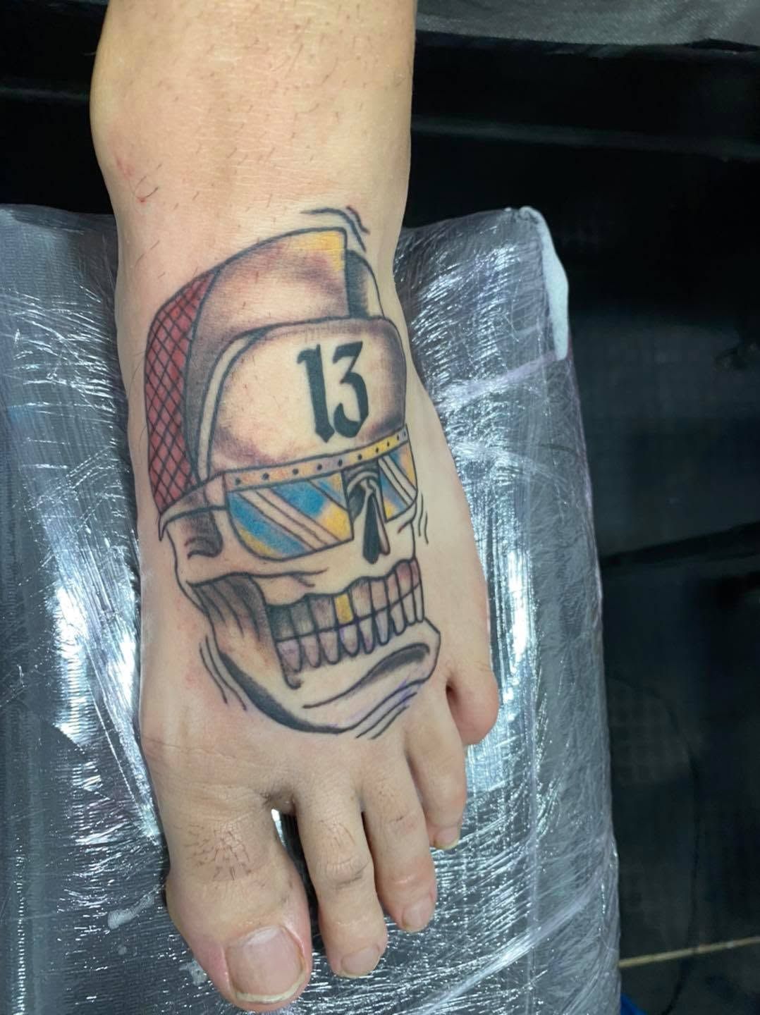 Pie con un tatuaje de calavera de colores y una gorra blanca marcada con el número «13» en el empeine
