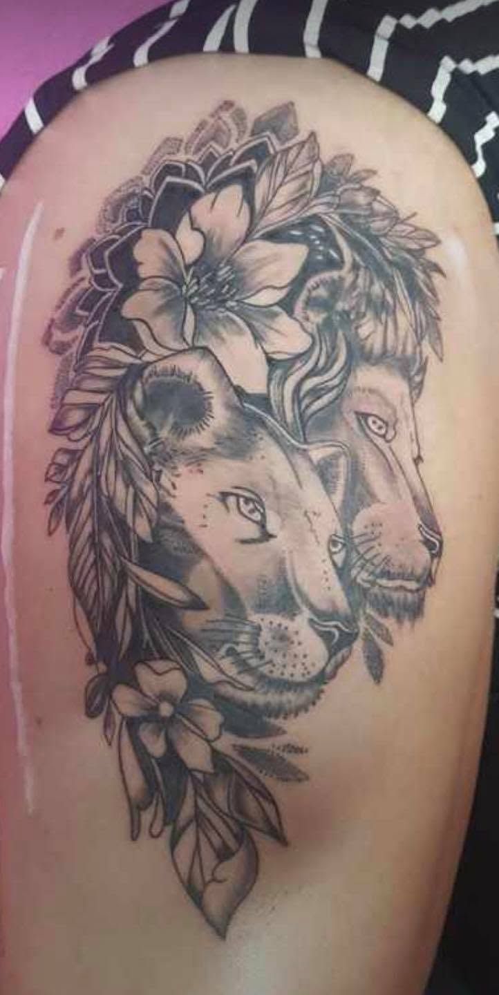 Tatuaje en negro y gris de un león con una corona floral sobre la piel, con el rostro estilizado de una mujer fusionado con el del león.