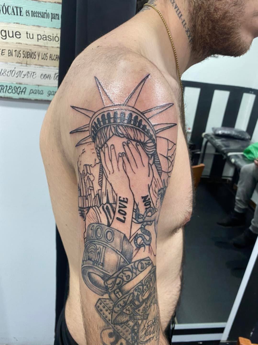Vista lateral del tatuaje en la parte superior del brazo de un hombre, con la Estatua de la Libertad y una mano cubriéndose la cara.
