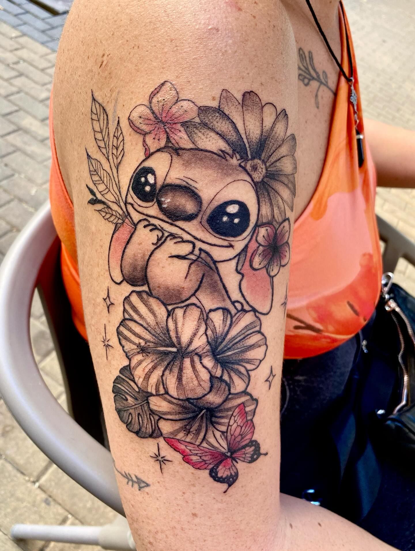 Tatuaje de estilo caricaturesco en la parte superior del brazo: un extraterrestre con flores, ojos grandes y una rama en la mano, en tinta negra y rosa