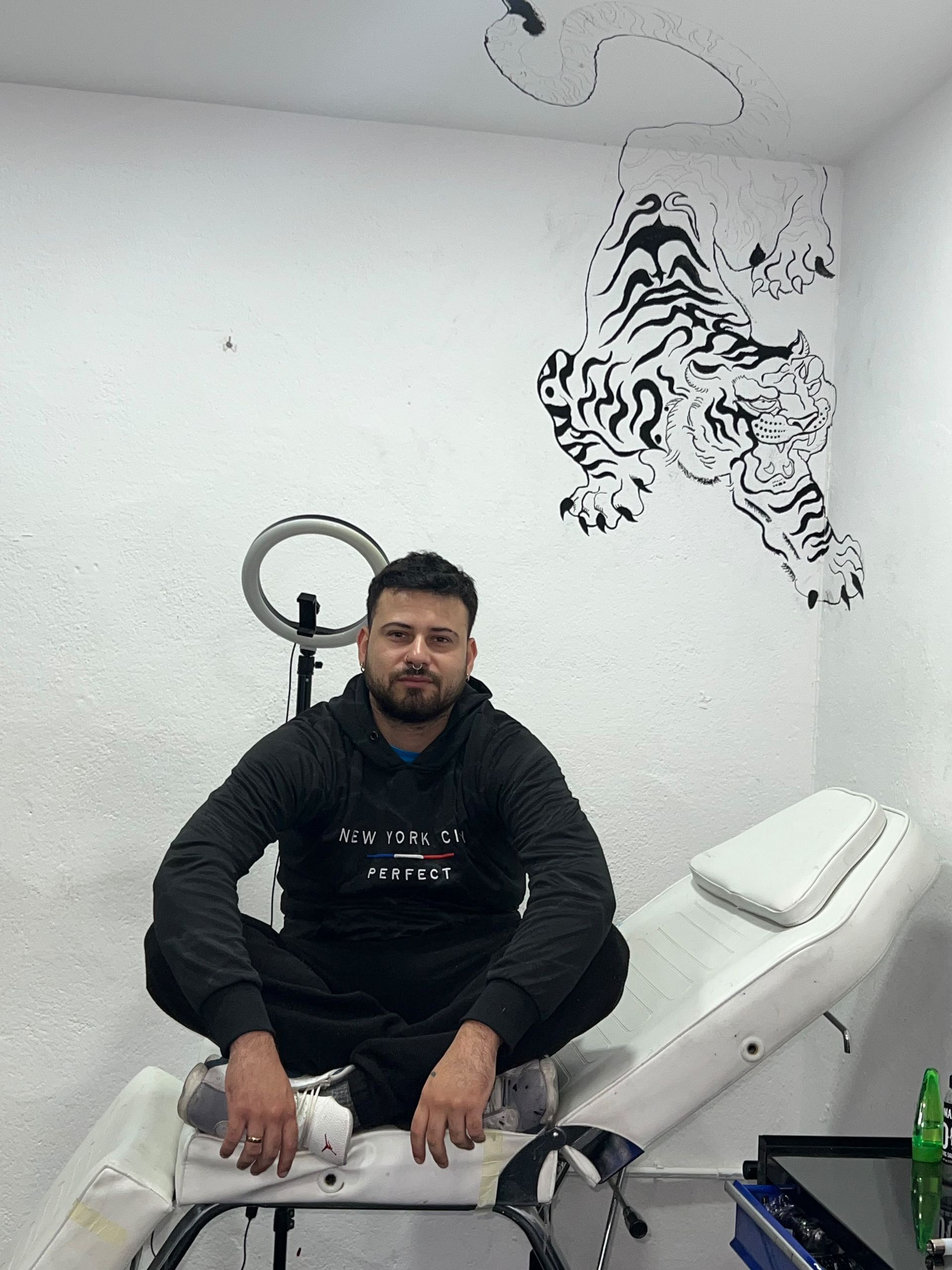Un hombre sentado en un sofá blanco en una habitación sencilla, con una obra de arte negra en la pared detrás de él.
