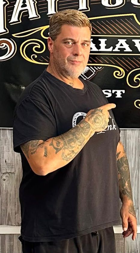 Hombre con camiseta negra y los brazos tatuados, de pie frente a un letrero decorativo en la pared.