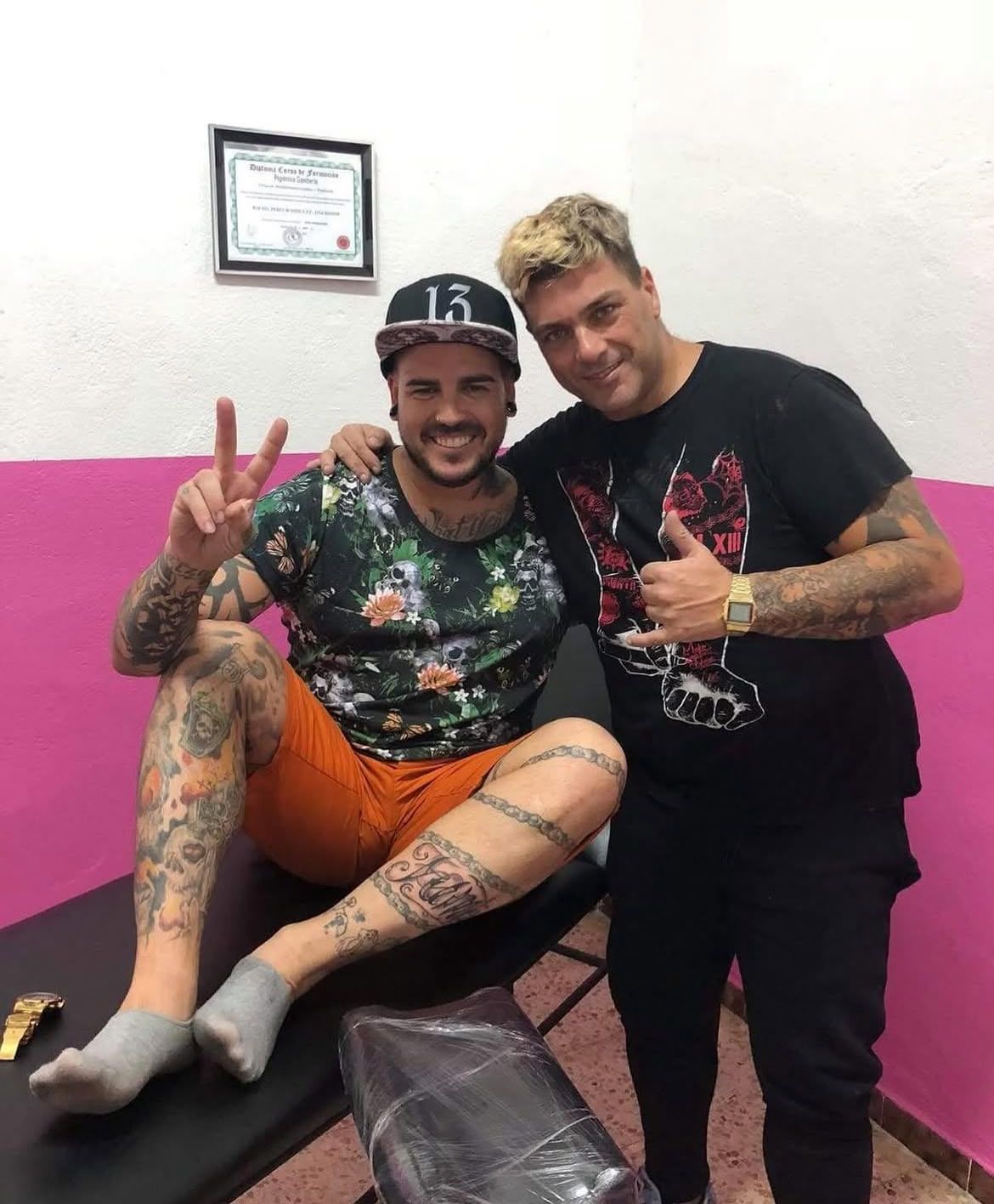 Dos personas sonrientes posan en un interior: una sentada con pantalones cortos naranjas y la otra de pie, ambas haciendo el signo de la paz.