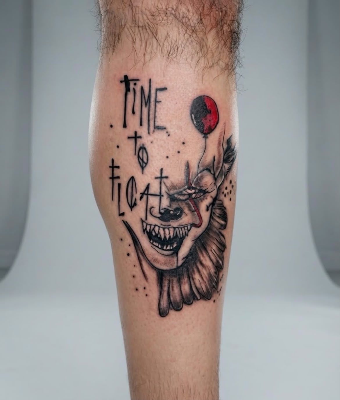 Pierna con un tatuaje de un payaso negro y rojo que dice «time to float», con una cara sonriente y un globo rojo